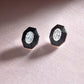 Pair of diamond stud earrings with black hexagonal settings on a pink fabric background