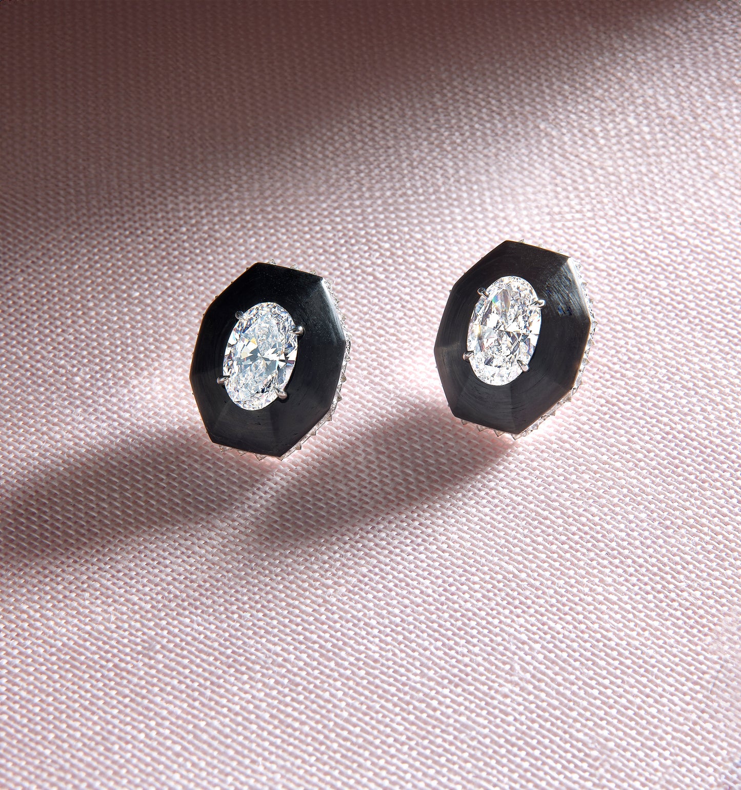 Pair of diamond stud earrings with black hexagonal settings on a pink fabric background